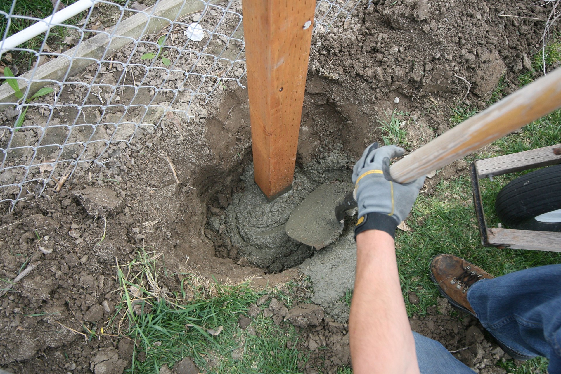 Worker installing a wooden fence post with concrete in a garden.